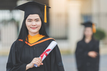 A young Asian woman university graduate in graduation gown and mortarboard holds a degree certificate stands in front of the university building after participating in college commencement