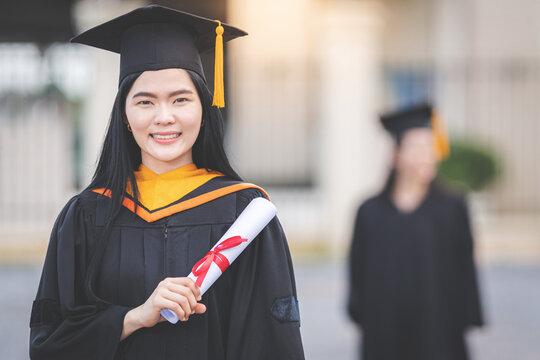 A young Asian woman university graduate in graduation gown and mortarboard holds a degree certificate stands in front of the university building after participating in college commencement