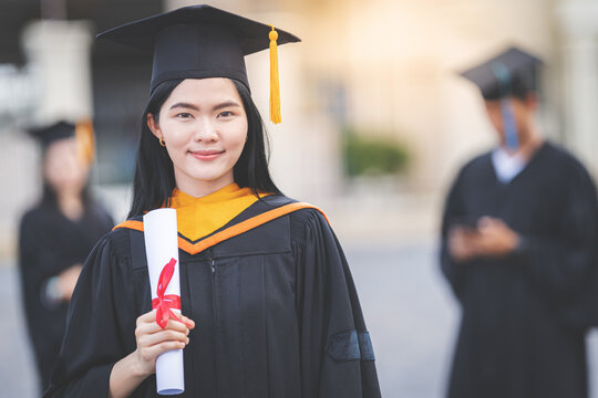 A young Asian woman university graduate in graduation gown and mortarboard holds a degree certificate stands in front of the university building after participating in college commencement