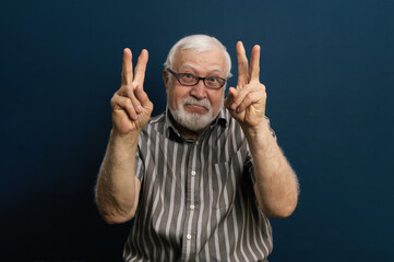 Fototapeta premium Charismatic gray-haired man in glasses shows two fingers, emotion in the frame, half-length portrait, blue background