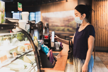 Woman customer with protective mask paying bill by cell phone in cafe.