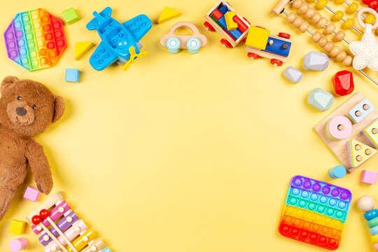 Baby kids toy background with teddy bear, wooden and musical toys, abacus, plane, pop it fidget toys and colorful blocks on yellow background. Top view, flat lay