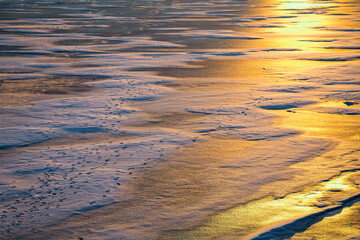 Winter landscape of frozen lake on sunset. Snow, ice and puddles. Many footprints on ice. It is dangerous to walk or fish on fragile ice in winter.