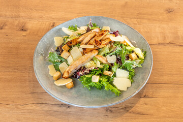 Caesar salad with pieces of grilled chicken breast with fried croutons with parmesan cheese flakes and assorted lettuce sprouts on a pearly gray plate