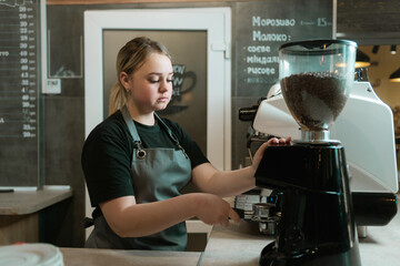 A barista grinds beans in a coffee grinder in a cafe and holds a portafilter. Sad girl waiter with...