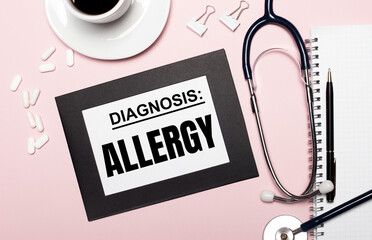 On a light pink background, a notebook with a pen, stethoscope, white pills, paper clips and a...