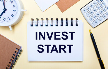On a light background, a white alarm clock, a calculator, a pen and a notebook with the text INVEST START. Flat lay