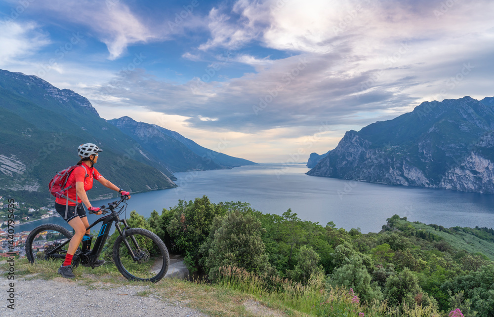 Wall mural nice senior woman with elctric mountain bike resting on Monte Brione and enjoying the awesome view over Garda Lake between Riva del Garda and Torbole