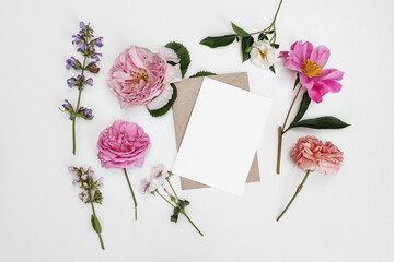 Floral summer stationery mockup. Blank greeting card with envelope. Garden flowers and herbs isolated on white table background in sunlight. Roses, sage, peony and geranium blooms. Flat lay, top view.