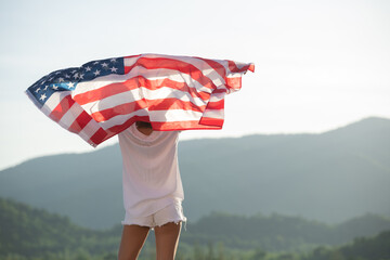 Back view happy young woman posing with USA national flag standing outdoors at sunset. Positive girl celebrating United States independence day. International day of democracy concept.