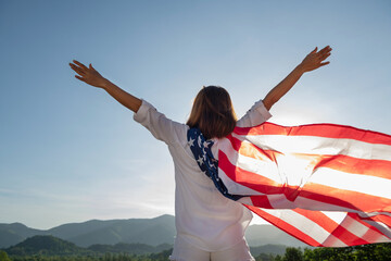 Back view happy young woman posing with USA national flag standing outdoors at sunset. Positive girl celebrating United States independence day. International day of democracy concept.