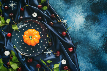 Halloween composition with a pumpkin on a plate, cherries, candle ends and decorative spiders on a blue satin draped fabric on a blue background with copy space. Helloween still life, flat lay