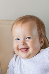 Fototapeta premium Cute caucasian infant girl ten months old sitting in baby chair and smiling on beige background. Closeup portrait of happy infant with blonde hair and blue eyes