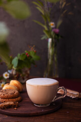 coffee with milk foam in a glass mug on a wooden table next to a bouquet of wildflowers and cookies
