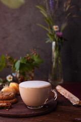 coffee with milk foam in a glass mug on a wooden table next to a bouquet of wildflowers and cookies