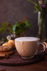 coffee with milk foam in a glass mug on a wooden table next to a bouquet of wildflowers and cookies
