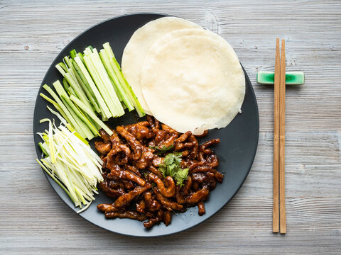 chinese cuisine - top view of pork in peking soy sauce with sliced scallion, cucumber and rice pancakes on black plate on wood table