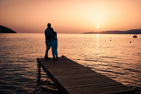 a couple seated on a wooden jetty, looking at colorful sunset on the sea, men, and women watching a sunset in Crete Greece Europe - Powered by Adobe