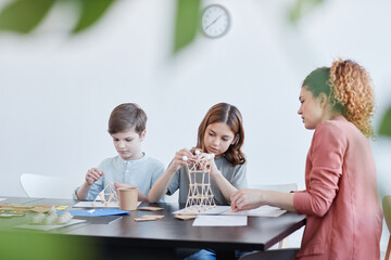 Side view portrait of female teacher helping children making wooden models during art and craft class in school, copy space