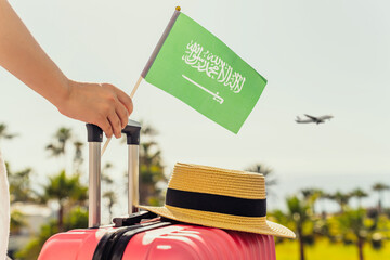 Woman with pink suitcase and Saudi Arabia flag standing on passengers ladder and getting out of airplane opposite sea coastline with palm trees.