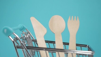 Wooden disposable recycled kitchen utensils in a small shopping cart. Free plastic concept. Wood spoon, fork and knife in a decorative supermarket trolley. Close up. Blue background