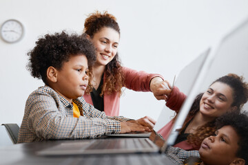 Side view portrait of young female teacher helping African-American boy using laptop during IT...