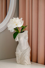 Conceptual image of a single peony flower in ceramic vase in shape of 150 BC era statue venus de milo on table with a mirror above on the wall. Close up shot of bedroom corner. Copy space for text.