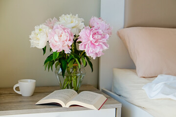 Close up shot of the nightstand with a book and bouquet of peonies in a glass vase near the unmade...