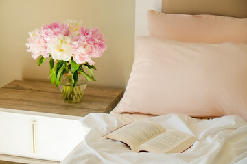 Close up shot of the nightstand with a book and bouquet of peonies in a glass vase near the unmade bed. Good morning concept. Bedroom full of natural light. Copy space for text, background, top view.