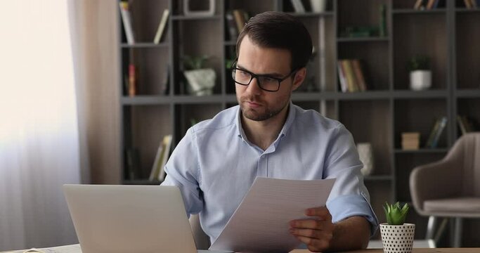 Serious businessman read contract papers learn formal document sit at workplace desk in modern office, do paperwork, comparing information looks at laptop, lawyer assistant attorney occupation concept