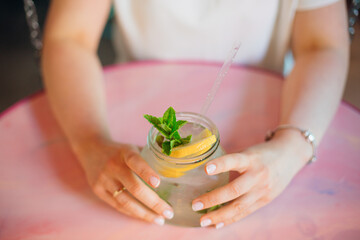 Young woman in summer sits at pink round table in cafe and hugs jug of deliciously cold lemonade. Travel and recreation. Serenity and relaxation. Refreshing drinks. Getting pleasure from life.