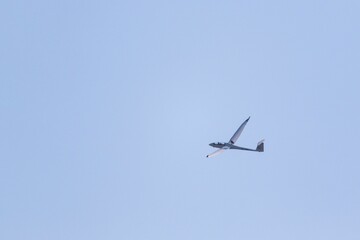 A portrait of a glider plane flying in the distance in a light blue sky. The plane is soaring...