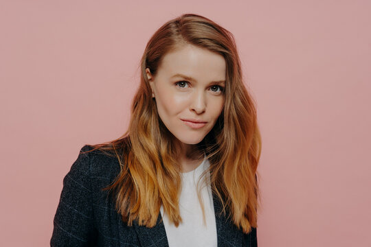 Attractive young woman with ginger hair in formal jacket posing against pink background