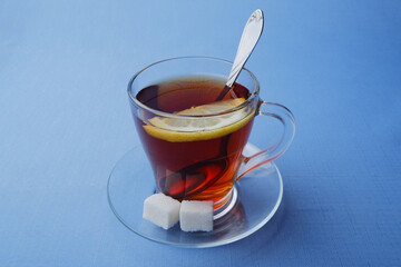a glass of tea with lemon and two pieces of sugar stands on a glass saucer on a blue background
