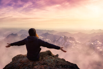Adventure Composite. Adventurous Man with Open Hands is taking in the moment on top of a mountain. Colorful Sunset or Sunrise Sky. Aerial Background Landscape from British Columbia, Canada.