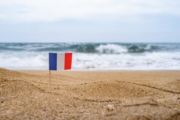 Flag of France in the form of a toothpick in the sand of beach opposite sea wave. Travel concept.