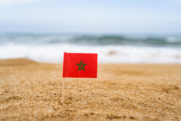 Flag of Morocco in the form of a toothpick in the sand of beach opposite sea wave. Travel concept