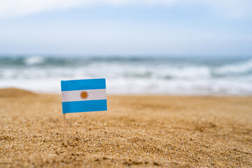 Fototapeta na wymiar Flag of Argentina in the form of a toothpick in the sand of beach opposite sea wave. Travel concept