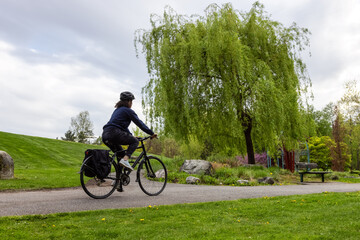 Naklejka premium Adult Caucasian Woman Riding a Bicycle on a path in a modern city park. Spring Evening. Taken in Hawthorne Rotary Park, Surrey, Vancouver, British Columbia, Canada.