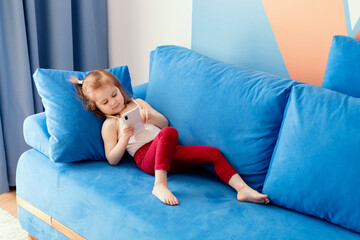 Caucasian little toddler girl wear casual clothes lying on blue coach in domestic room, holding...