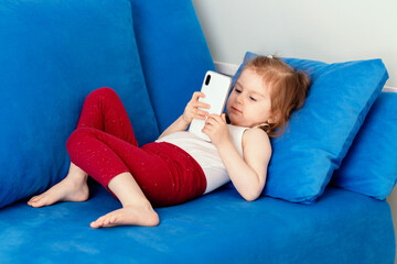 Thoughtful little toddler girl wear casual clothes lying on blue coach in domestic room, holding smartphone and watching cartoons.