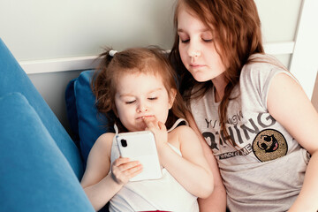 Two sisters lying on blue sofa at domestic room and watching cartoons films on smartphone.