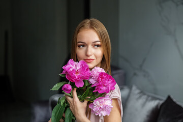 A beautiful girl with long blonde hair in a modern interior holds a bouquet of pink peonies in her hands, a romantic girl