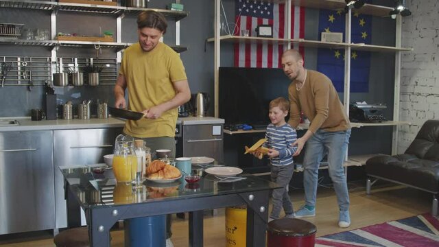 Slowmo medium shot of cheerful man serving breakfast while his male partner and playful school-age boy sitting down to eat