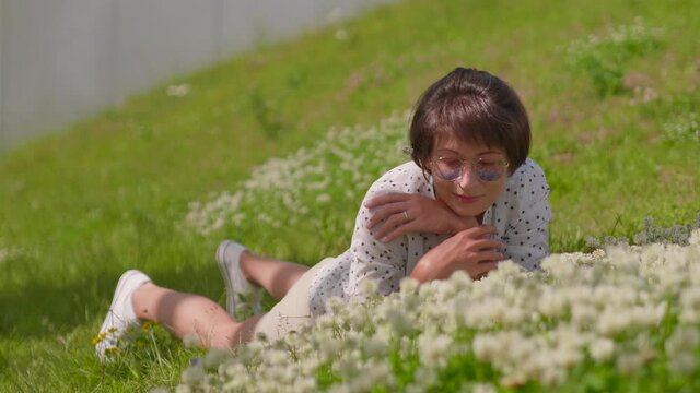 Woman with colorful sunglasses lays on lawn in urban park and enjoys flowers. Summer heat in town.