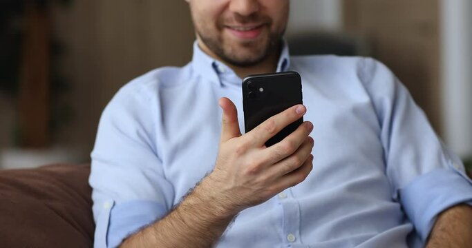 Close up cropped view young smiling man sit on couch with smart phone device, texting message, send sms using modern wireless tech, e-commerce client purchasing via e-store mobile application concept
