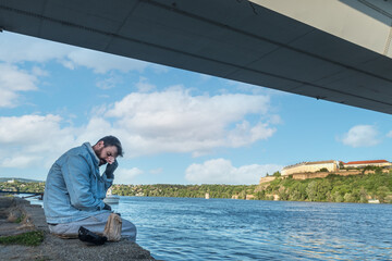 Young sick desperate depressed alcoholic homeless man sitting on the dockyard under the bridge drinking beer and feeling miserable and hopeless because of his sad and lonely life, social documentary 