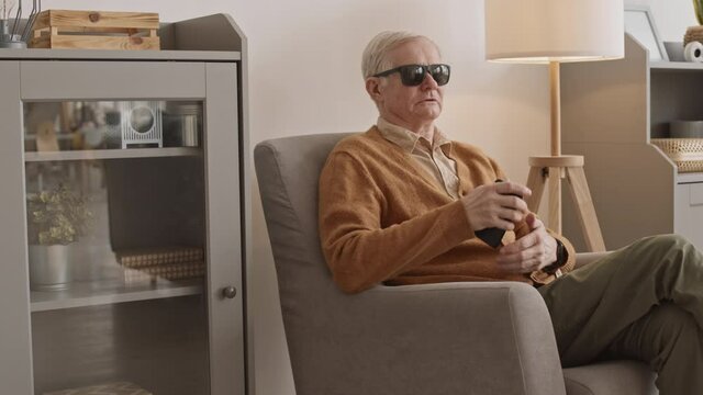 Medium slowmo of blind senior man in black eyeglasses using voice assistant on his smartphone while sitting in chair at homeMedium slowmo of blind senior man in black eyeglasses using voice assistant 