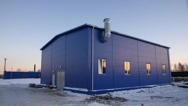 Large modern workshop building with metal chimney on snowy ground under clear blue sky at sunset in winter evening