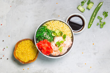 Poke, one of the main dishes of Hawaiian cuisine. Bowl with bulgur, shrimp, chuka salad. On a gray background with sauce, grits in a wooden bowl and chili pepper. Horizontal, overhead